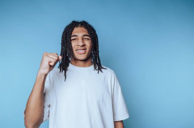 Genç bir adam açık mavi bir zemine karşı duruyor, güven ve kararlılık jesti olarak yumruğunu kaldırıyor. Uzun kıvırcık saçları neşeli bir ifade çiziyor, pozitifliği yansıtıyor..