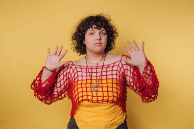 Bir insan, parlak sarı bir arkaplan önünde ellerini kaldırarak güvenle durur. Sarı bir tişörtün üzerine kırmızı ağlı bir bluz giyiyorlar. Benzersiz bir stil ve kişilik ifadesi sergiliyorlar..