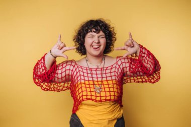 Kıvırcık saçlı genç bir kadın canlı sarı bir arkaplanda poz verirken neşeli bir ifade sergiliyor. Sarı bir tişörtün üzerine kırmızı bir örtü giyer ve eğlenceli bir hava yayar..