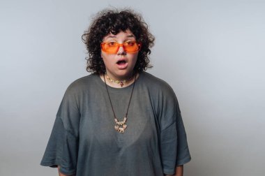 Kıvırcık saçlı ve turuncu güneş gözlüklü bir kadın minimalist stüdyo arkaplanına şaşırmış görünüyor. Gevşek gri bir gömlek ve boncuklu bir kolye giyiyor..