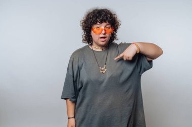 Kıvırcık saçlı ve turuncu güneş gözlüklü şahıs kendine güvenen bir duruş sergiliyor. Kendilerini gri, büyük bir tişörtle gösteriyorlar. Stüdyo ortamında eğlenceli ve rahat bir hava sergiliyorlar..