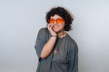 Son moda turuncu güneş gözlüğü takan ve gevşek gri tişörtlü genç bir kadın açık bir arka planda duruyor, gülümsüyor ve eli yüzünde şakacı bir şekilde poz veriyor..