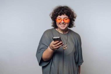 Kıvırcık saçlı ve turuncu güneş gözlüklü bireyler akıllı telefonlarına göz atmaktan zevk alırlar. Kapalı bir alanda, tarafsız bir arka planda neşeli bir titreşim yayarlar..