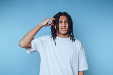 Genç bir adam açık mavi bir arka fonun önünde duruyor, düşünceli bir jestle kafasını kaşıyor, rahat bir ortamda şaşkınlığı ya da merakı gösteriyor, muhtemelen bir soruyu yansıtıyor..