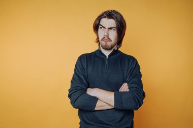 Genç bir adam kollarını kavuşturmuş, rahatsız edici bir bakış atıyor. Parlak turuncu arka plan o ana enerji katıyor, ciddi davranışlarını vurguluyor..