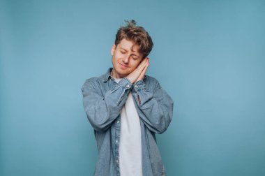 Kıvırcık saçlı genç bir adam uyku pozunu taklit ederken gülümsüyor, neşeli bir ruh hali sergiliyor. Yumuşak mavi arka plan neşeli atmosferi arttırır, onu davetkar ve sıcak yapar..