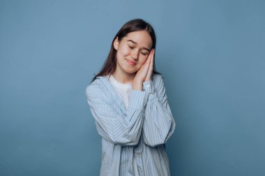 Genç bir kadın açık mavi bir duvarın önünde duruyor, gözlerini kapıyor ve başını huzurlu bir jest yaparak nazikçe gülümsüyor. Rahatlamış tavrı mutluluğu taşıyor..