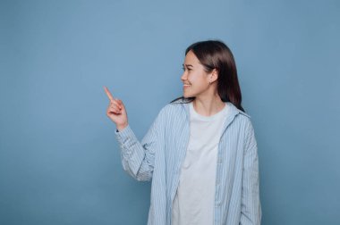 Genç bir kadın açık mavi bir arka plana karşı duruyor, çizgili bir gömlek ve sade beyaz bir tişört giyiyor. Gülümsüyor ve sağını işaret ediyor, coşku ve nişanı iletiyor..