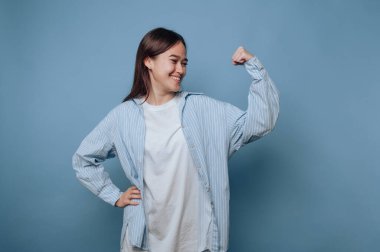 Genç bir kadın mavi bir arka plana karşı gururla duruyor, gücünü göstermek için kolunu esnetiyor. Beyaz bir tişörtün üzerine açık mavi çizgili bir gömlek giyer, kendine güvenle gülümser..