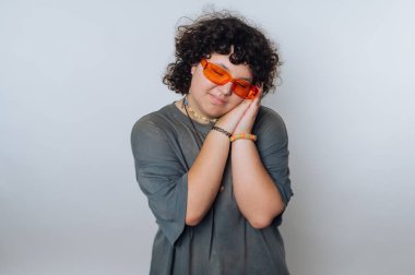 Kıvırcık saçlı bir genç günlük kıyafet giyer ve neşeli poz verirken parlak turuncu güneş gözlüğü takar. Stüdyo geçmişi basit ve tarafsız..