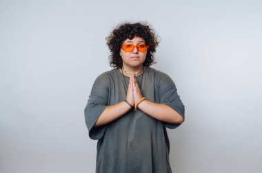 Kıvırcık saçlı biri, bir teşekkür jesti ile elini sıkarak ayakta duruyor. Gündüz arkaplanında turuncu gözlüklü eşsiz bir stil sergiliyor..