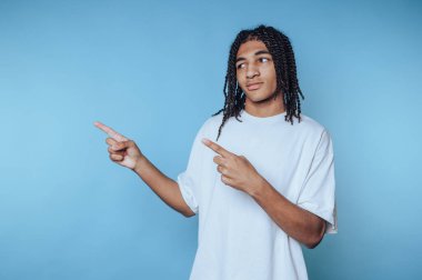 Genç bir adam, beyaz gömlek ve örgü takmış, kendinden emin bir şekilde ayakta duruyor. Neşeli bir şekilde iki yönü işaret ediyor. Hevesini ve bağlılığını dostça gösteriyor..