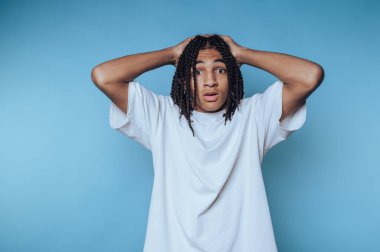 Genç bir birey elleri başlarının üstünde duruyor, şok veya inançsızlık ifadesi sergiliyor. Sade mavi arkaplan, ifadelerini ve günlük kıyafetlerini vurguluyor..