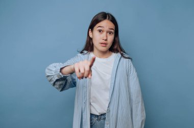 Genç bir kadın rahat bir şekilde durup parmağını izleyiciye doğrultuyor. Sıradan bir kıyafet giyiyor ve ciddi bir ifade takınıyor. Sade mavi bir alanda ilgi çekici bir an yaratıyor..