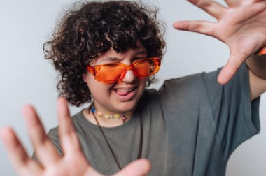 Kıvırcık saçlı genç bir birey neşeli el hareketleriyle neşeli bir kişilik sergiliyor, parlak turuncu güneş gözlüğü takıyor ve stüdyo ortamında günlük bir gömlek giyiyor..