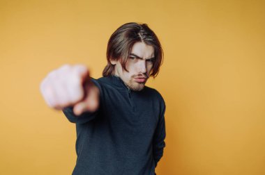 Uzun saçlı genç bir adam, parlak sarı bir duvara yaslanmış parmağını göstererek özgüvenle hareket eder. Azim ve tavır karışımı bir şey yayıyor..