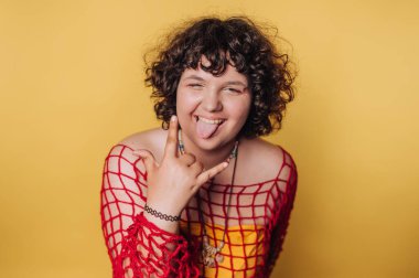 Kıvırcık saçlı genç bir birey el hareketi yaparken şakacı bir şekilde dilini uzatıyor. Parlak sarı arkaplan, o anın canlı atmosferini arttırır..