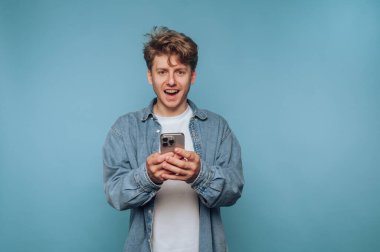 Kot ceket ve beyaz gömlek giyen genç bir adam akıllı telefon tutarken gülümsüyor. Neşeli ve meşgul görünüyor, mavi bir arkaplanın önünde duruyor..