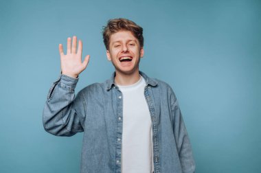 Açık kahverengi saçlı, kot gömlek giyen ve elini sallarken gülümseyen beyaz tişörtlü genç bir adam. Neşeli ifade ve parlak mavi zemin canlandırıcı bir atmosfer yaratır..