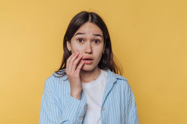 Uzun siyah saçlı genç bir kadın yüzüne dokunurken şaşırmış bir ifade gösterir. Parlak sarı bir arkaplanın önünde dikiliyor, rahat giyiniyor..