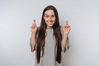 Uzun, düz saçlı genç bir kadın açık bir arka planda duruyor. O, umutlarını ve beklentilerini neşeli bir şekilde ifade ederken, parmaklarını çaprazlarken geniş bir şekilde gülümser..