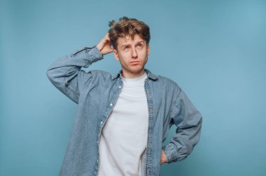 Genç bir adam, beyaz bir gömleğin üzerine kot ceket giymiş, dalgın bir ifadeyle ayakta duruyor. Düşüncelere dalmış gibi görünüyor, içsel bir ruh haliyle gündelik tarzını birleştiriyor..