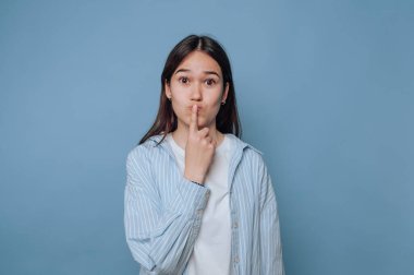 Genç bir kadın neşeli bir ifadeyle ayakta duruyor, parmakları dudaklarında sanki sessizlik istiyormuş gibi. Rahat bir kıyafet giyer. Doğrudan seyirciye bakar. Saf mavi bir arkaplan..