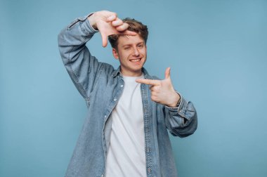 Genç bir adam açık mavi bir zemine karşı duruyor ve elleriyle bir çerçeve oluşturuyor. Beyaz bir gömleğin üzerine kot ceket giyiyor, yüzünde neşeli bir ruh var..