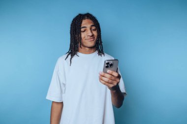 Örgülü saçları olan genç bir adam, elinde akıllı telefonu tutarken gülümseyen, rahat ve memnun bir tavır sergileyen, saf mavi bir fon karşısında duruyor..