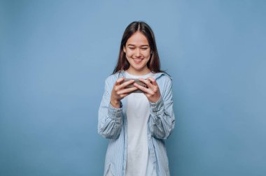 Genç bir kadın elinde akıllı telefonu tutarken gülümsüyor. O, açık mavi bir gömlek giyer, koyu mavi bir arka plan önünde eğlenir..