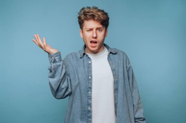 Genç bir adam açık mavi bir arka plana karşı duruyor, kot gömlek ve beyaz tişört giyiyor. Kafası karışmış görünüyor, bir elini kaldırırken göz kırpıyor, inançsızlığını dile getiriyor..