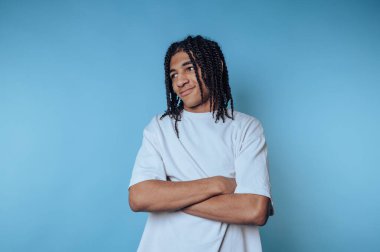 Kollarını kavuşturmuş genç bir adam rahat ama kendinden emin bir tavır sergiliyor. Saçlarını örüyor ve kameraya bakarken hafifçe gülümsüyor. Makarna mavisi bir arkaplana yaslanıyor..