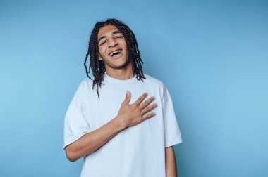 Saçları örgülü genç bir adam parlak mavi bir arka planda dururken yürekten güler. Elini kalbine koyar, gerçek mutluluğu ve sıcaklığı gösterir..