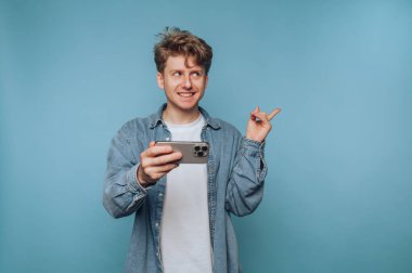 Neşeli genç bir adam açık mavi bir duvara karşı duruyor, akıllı telefonunu tutarak selfie çekiyor. Günlük kıyafetleri ve gülüşü dostane bir ortam yaratır..