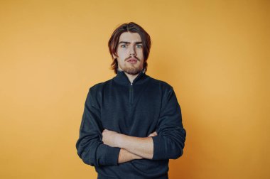 Genç bir adam, kollarını parlak sarı bir arka plana karşı kavuşturarak güvenle ayakta duruyor. Onun ciddi ifadesi neşeli renkle çelişiyor, çarpıcı bir görsel etki yaratıyor..