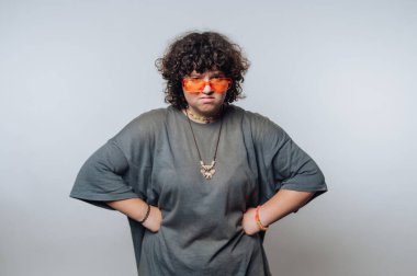 Kıvırcık saçlı ve güneş gözlüklü biri stüdyo ortamında güven içinde duruyor. Kişi bol giysi ve mücevher giyer, cesur bir tarz ve tavır sergiler..