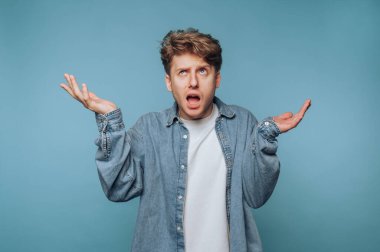 Genç bir adam ellerini havaya kaldırarak şaşkınlığını gösteriyor. Saçları dağınık ve beyaz bir tişörtün üstüne kot ceket giymiş..