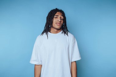 Rahat bir duruşu olan genç bir adam, basit beyaz bir tişört giyiyor. Örgülü saçı, parlak mavi bir arkaplana karşı düşünceli ifadesini tamamlıyor..