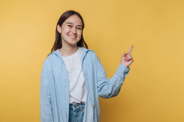 Genç bir kadın, parlak bir gülümsemeyle güvenle ayakta duruyor, sağ eliyle yukarıya bakıyor. Açık mavi çizgili bir gömlek ve kot giyiyor, neşeli sarı bir fona karşı..