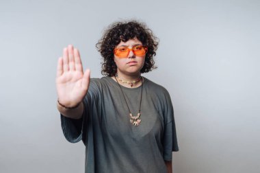 Kıvırcık saçlı, turuncu güneş gözlüklü ve gri tişörtlü biri, düz bir arkaplanda el kol hareketi yapıyor..