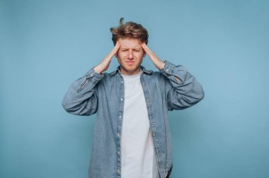 Kot gömlekli ve beyaz tişörtlü genç bir adam mavi bir arka plana karşı duruyor, iki eliyle başını tutuyor ve rahatsızlık veya stresi ifade ediyor..