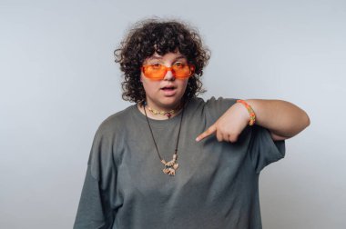 Kıvırcık saçlı, turuncu güneş gözlüklü ve kolyesini gösteren biri..