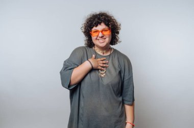 Kıvırcık saçlı, turuncu güneş gözlüğü takan ve gri tişörtlü bir adam göğsünde, düz bir arkaplanda sımsıkı gülümsüyor..