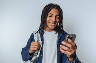 Saçları örgülü genç adam akıllı telefona bakarken gülümsüyor, sırt çantası taşıyor, kot ceket giyiyor ve beyaz tişört giyiyor, düz bir arkaplanda duruyor..