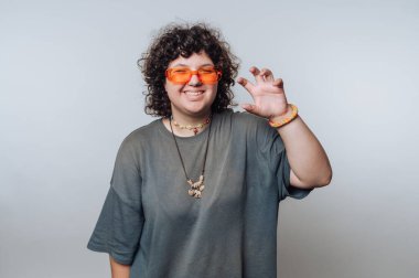 Kıvırcık saçlı, turuncu güneş gözlüğü takan ve gri tişörtlü, sade bir arka plana karşı şakacı bir pençe hareketi yapan biri..