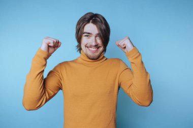 Hardal süveter giyen genç bir adam mavi arka planda yumruklarını kaldırarak kutluyor..