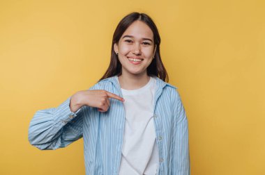 Genç bir kadın gülümsüyor ve sarı arka planda kendini işaret ediyor..