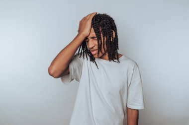 Beyaz tişörtlü, saçları örgülü genç bir adam. Kafasını düz bir arkaplanda hayal kırıklığı içinde tutuyor..