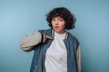 Kıvırcık saçlı, kot ceket giyen ve mavi arka planda beyaz gömlekli biri, kendini şaşırmış bir ifadeyle gösteriyor..