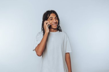 Rastalı genç adam akıllı bir telefonla konuşuyor, beyaz bir tişört giyiyor, düz beyaz bir arka plana karşı duruyor..